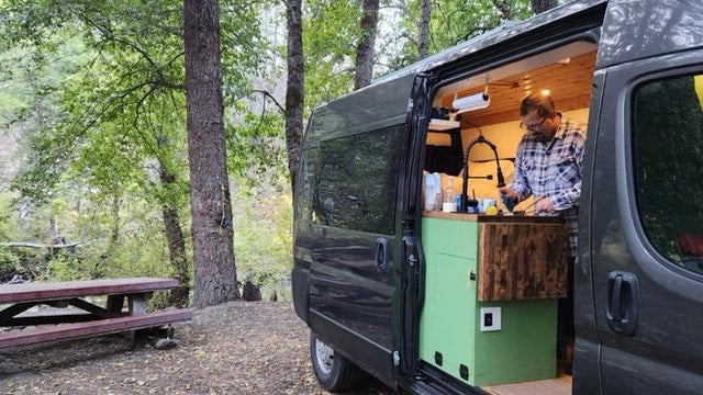 Man prepares food in an RV rental camper van's custom kitchen, parked at a wooded campsite with a picnic table. — photo 8