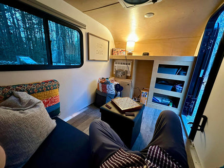 Serene RV rental interior: person relaxing on sofa, forest view, board game on ottoman, "Our Nest" sign.