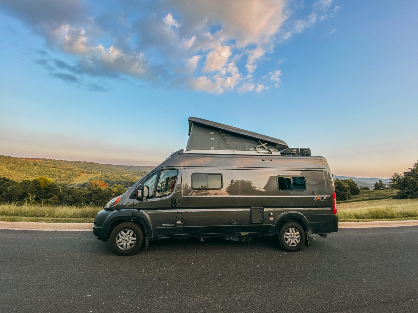 Grey RV rental (Winnebago Solis) with pop-top, parked on a road overlooking green hills under a blue sky. Ideal for van life adventure.