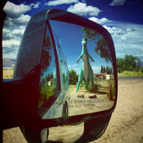 A huge green dinosaur statue eerily close in an RV rental mirror, a classic roadside surprise. Perfect for travel exploration!