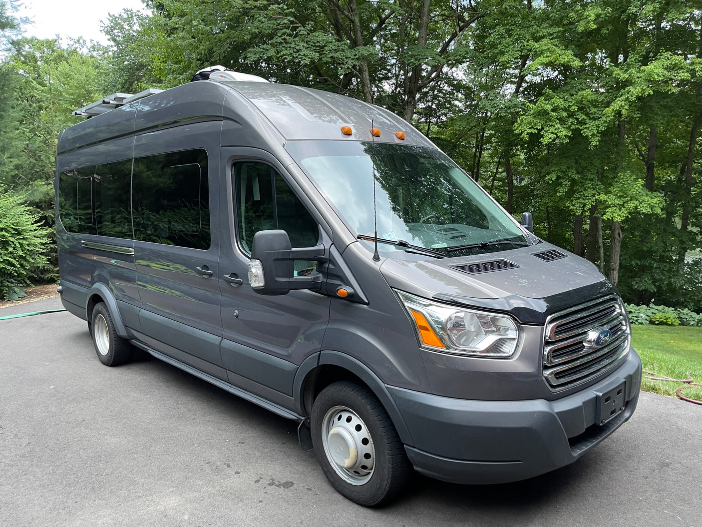 Gray Ford Transit rv rental van with solar panels & AC unit, parked outdoors among green trees.