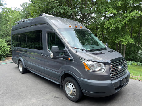 Gray Ford Transit rv rental van with solar panels & AC unit, parked outdoors among green trees.