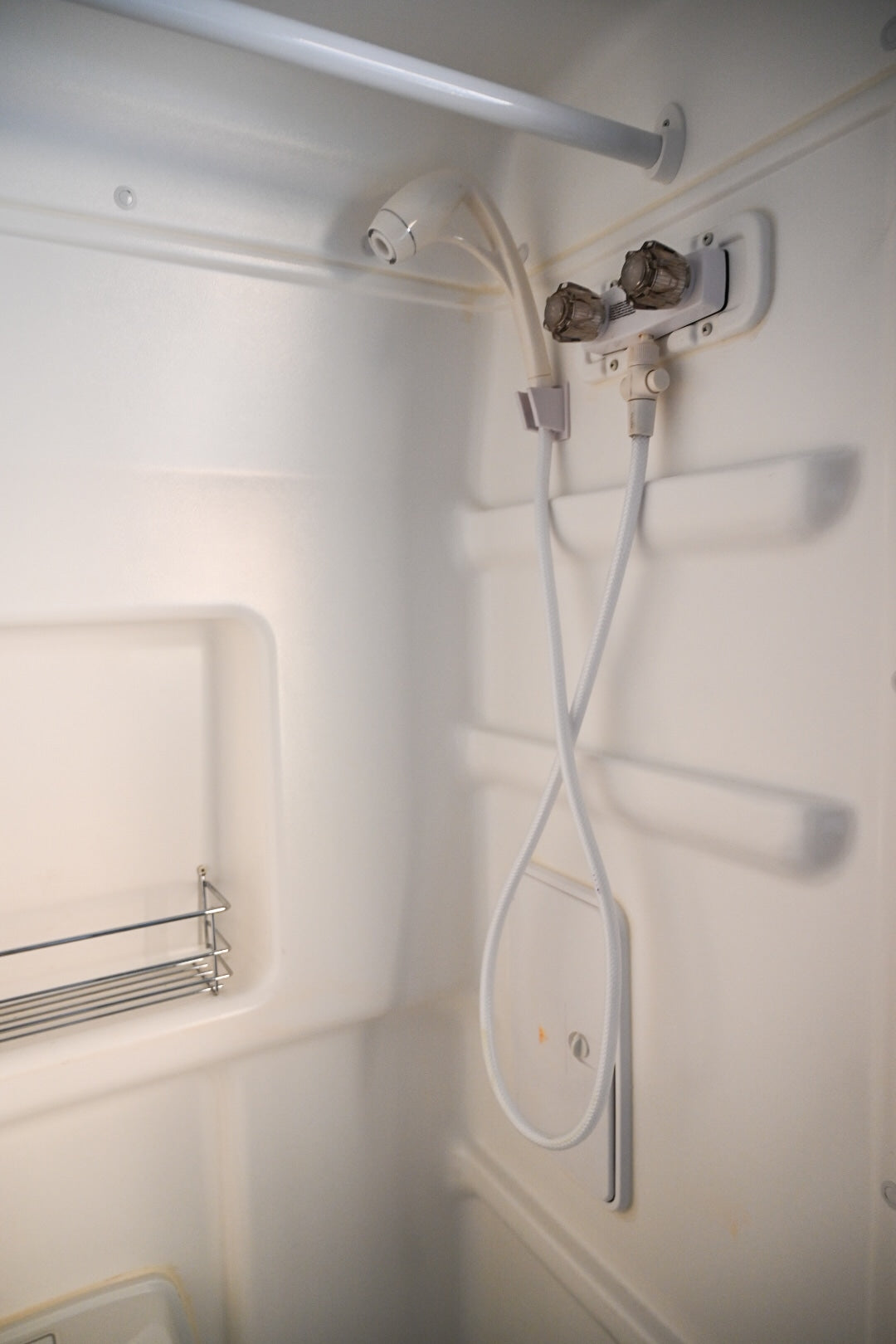Inside an RV rental shower: white molded stall with handheld shower, water controls, and a wire rack for essentials.