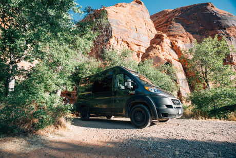 Dark green RV rental camper van parked on a dirt road by large red rock desert cliffs and green trees.