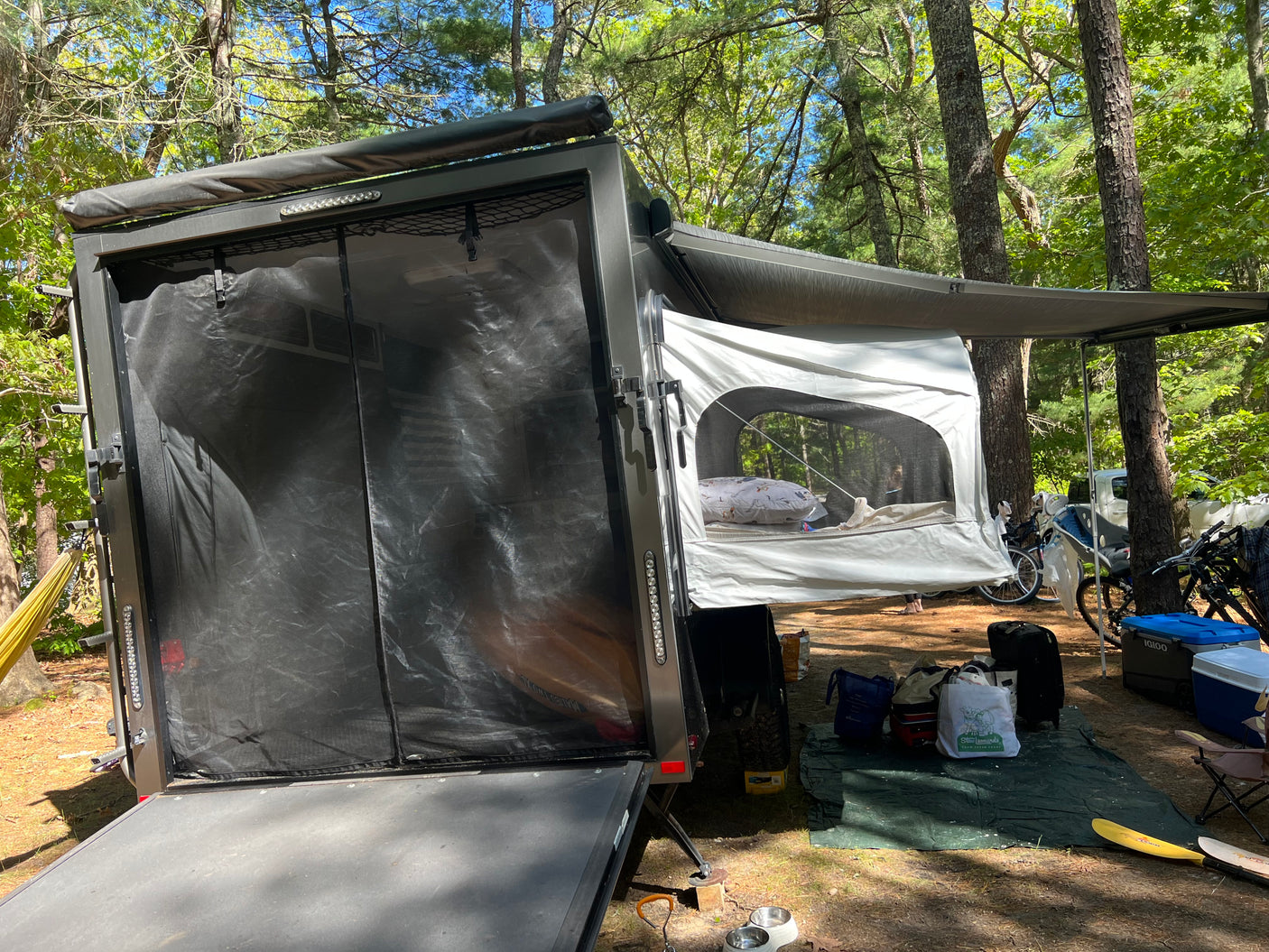 RV rental camping setup: a toy hauler with open ramp, pop-out bed, awning, bikes & coolers in a wooded campsite.