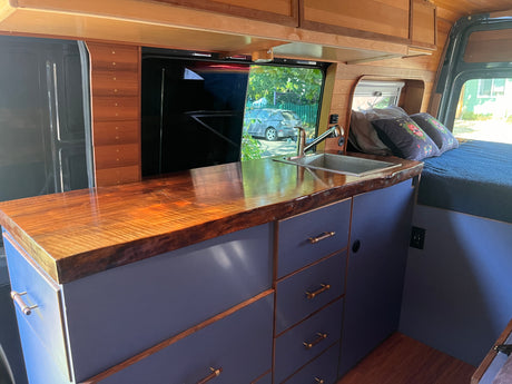 Inside a stylish RV rental: custom live-edge wood kitchen counter, sink, modern blue cabinets, and a comfy bed.