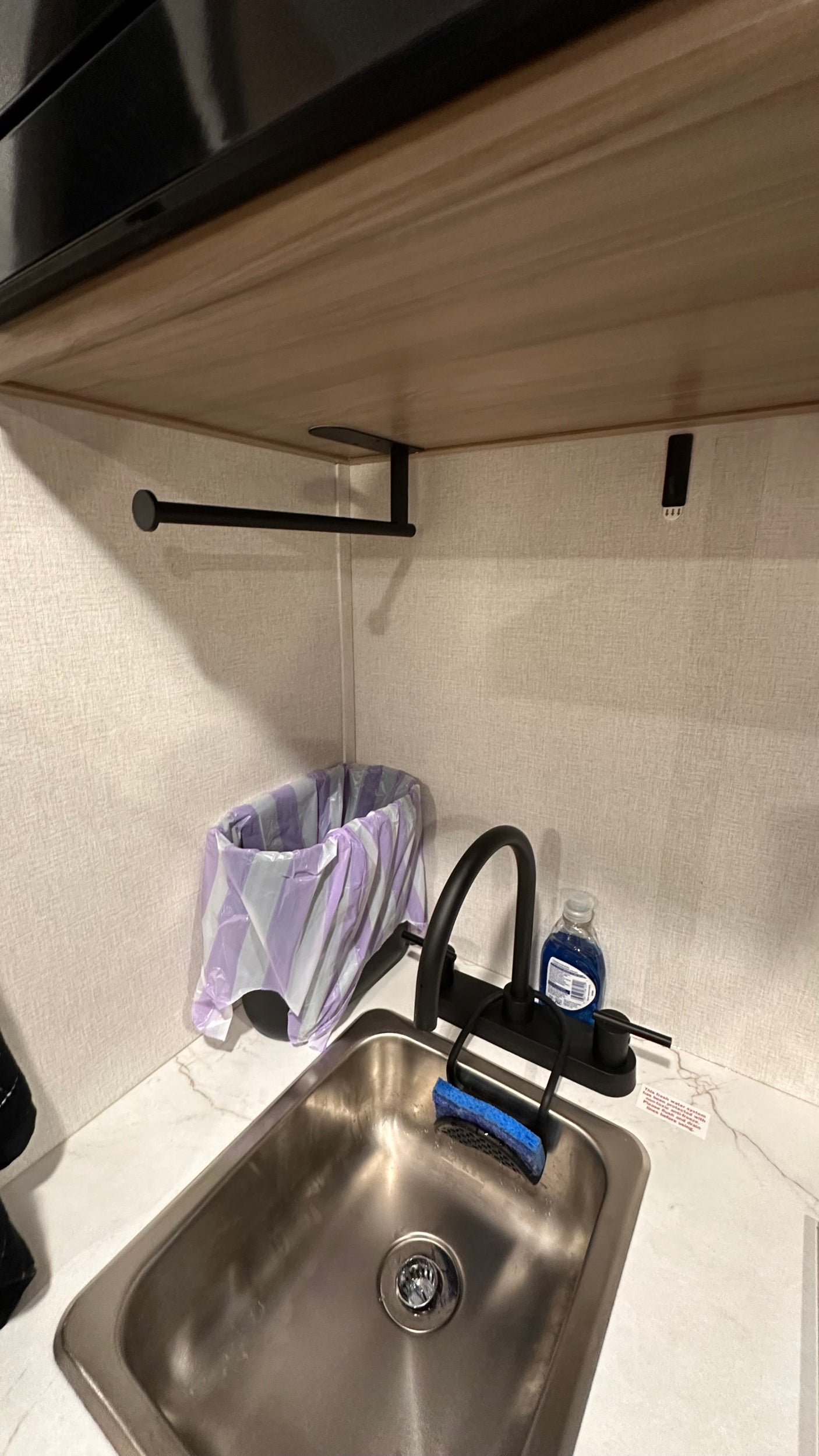 Compact RV rental kitchen area featuring a stainless steel sink, black faucet, dish soap, sponge, and trash can.