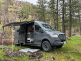 Spacious gray RV rental van with awning extended at a lush, rocky forest campsite, ideal for nature trips.