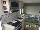 Compact RV rental interior: view of the kitchenette with a stove, microwave, and decorative backsplash, plus a bed area.