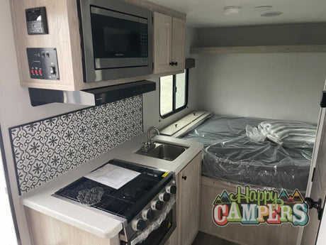 Compact RV rental interior: view of the kitchenette with a stove, microwave, and decorative backsplash, plus a bed area.