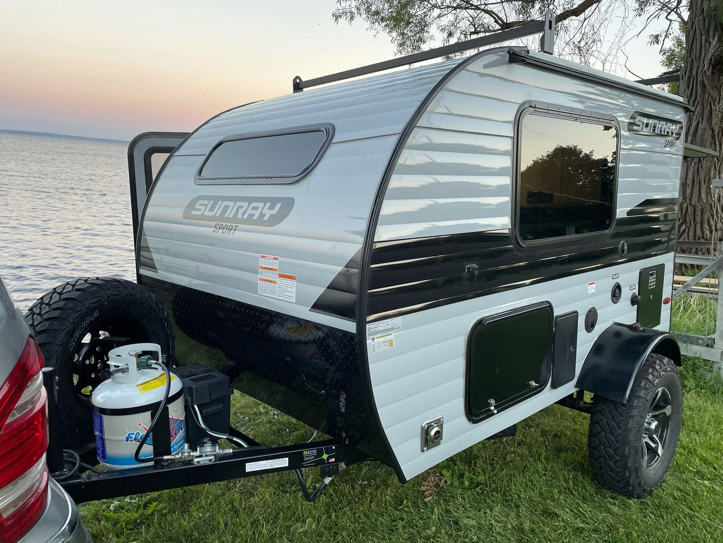 A light gray Sunray Sport travel trailer with off-road tires, perfect for an RV rental for lakeside camping at dusk.