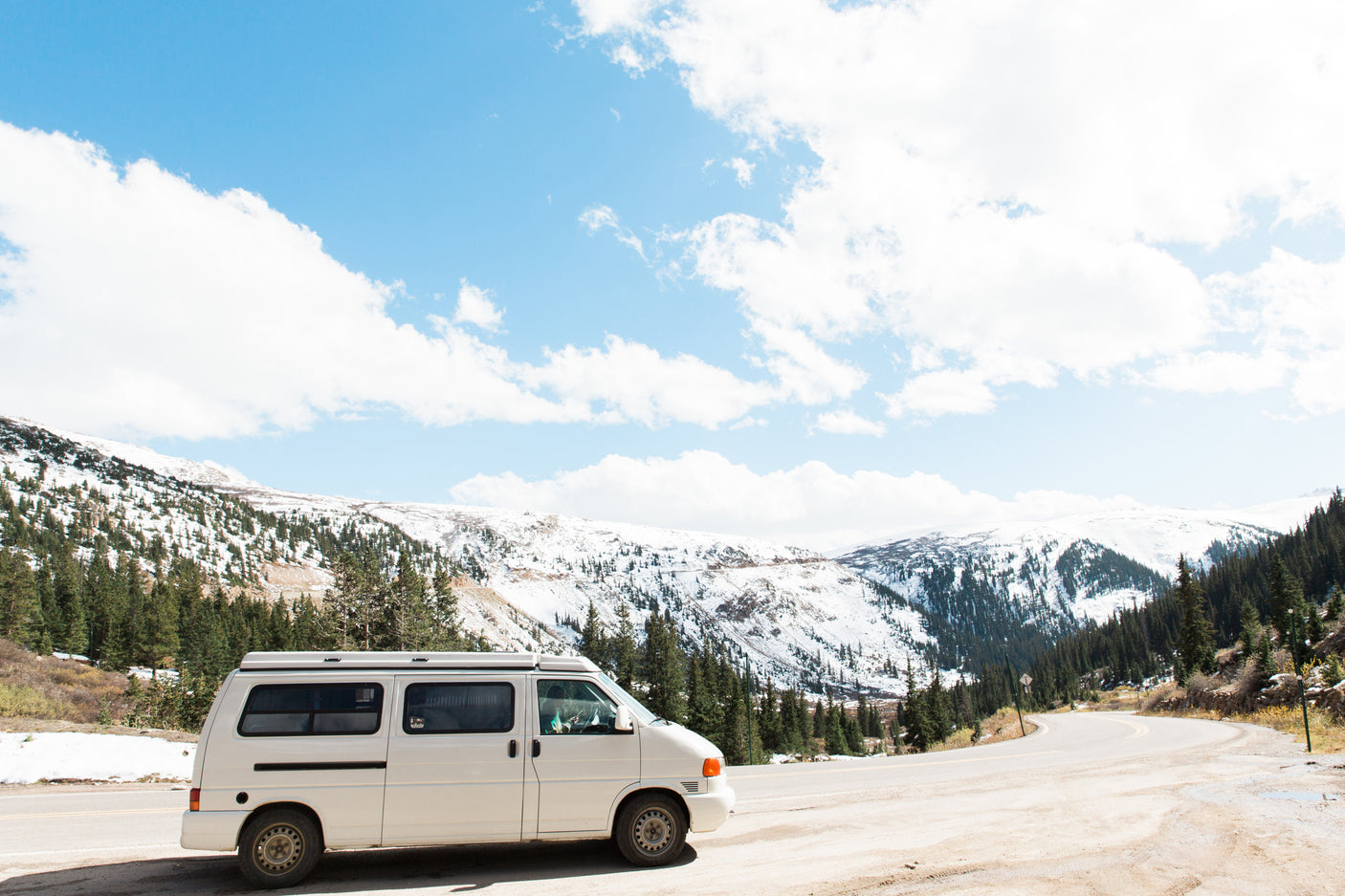 RV rental parked on a scenic mountain road with snow-capped peaks and evergreen trees under a blue sky.