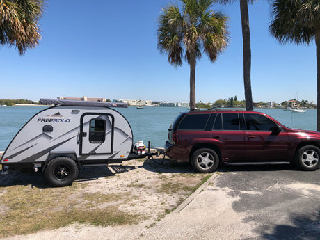 A grey Freesolo teardrop RV rental hitched to a red SUV, parked by a sunny waterfront with palm trees and boats in the distance.