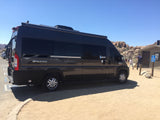 Dark gray RV rental camper van with awning & bike rack parked in a sunny, rocky desert landscape near restrooms.