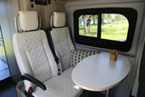 Bright RV rental interior featuring two cream-colored seats with textured patterns, a small table, and a window with a scenic park view.