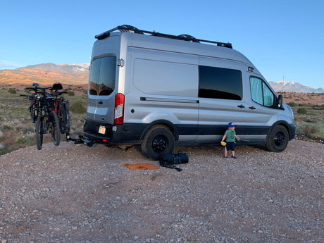 An RV rental silver camper van with bikes, parked in a stunning desert. A young child plays nearby.