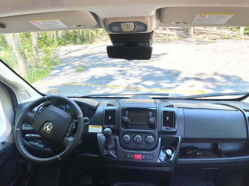 RV rental cockpit view: dashboard, steering wheel, and front windshield looking out to a wooded road.