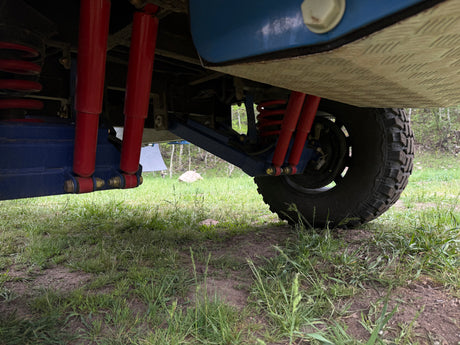 View of a rugged RV rental's undercarriage: red shocks, coil springs, blue suspension, and an aggressive off-road tire on dirt.