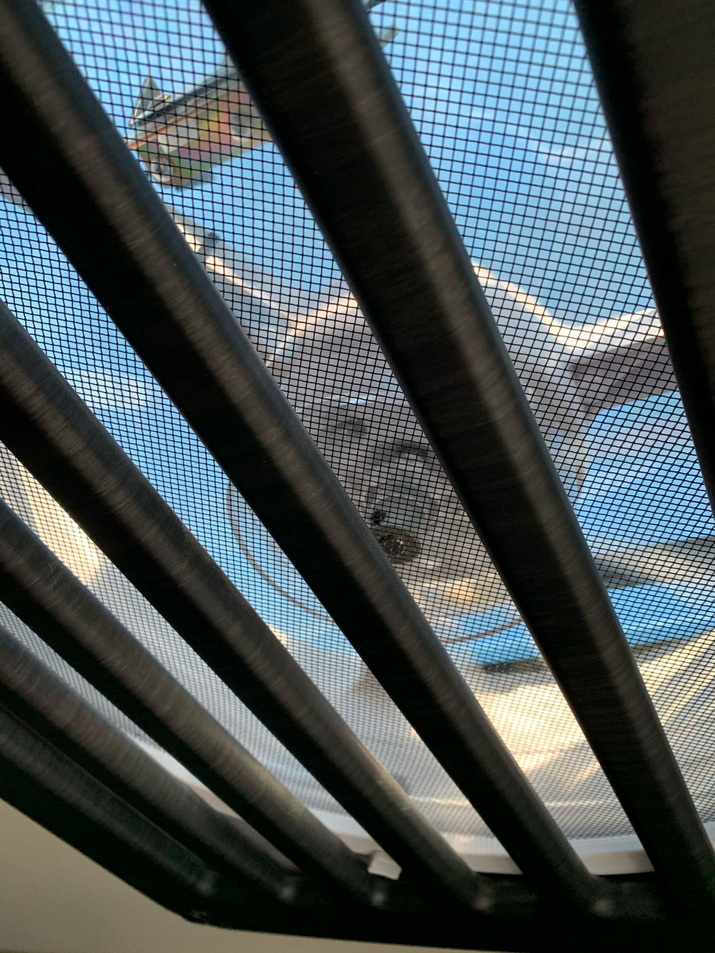 View from an RV rental looking up through dark blinds and a screen at a colorful kite and blue sky.