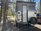 This pop-up RV rental is set up in a pine forest, showing its rear with a spare tire and steps for camping adventures.