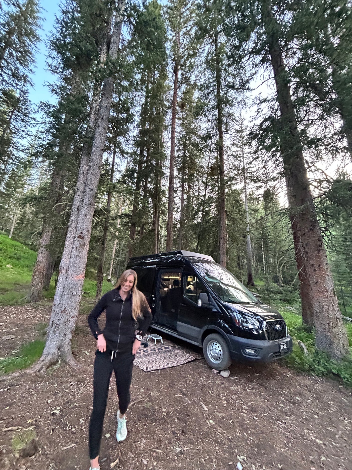 Forest camping with an rv rental. A smiling woman in a black tracksuit stands beside her black camper van among tall trees.