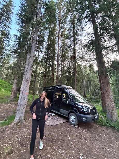 Forest camping with an rv rental. A smiling woman in a black tracksuit stands beside her black camper van among tall trees.