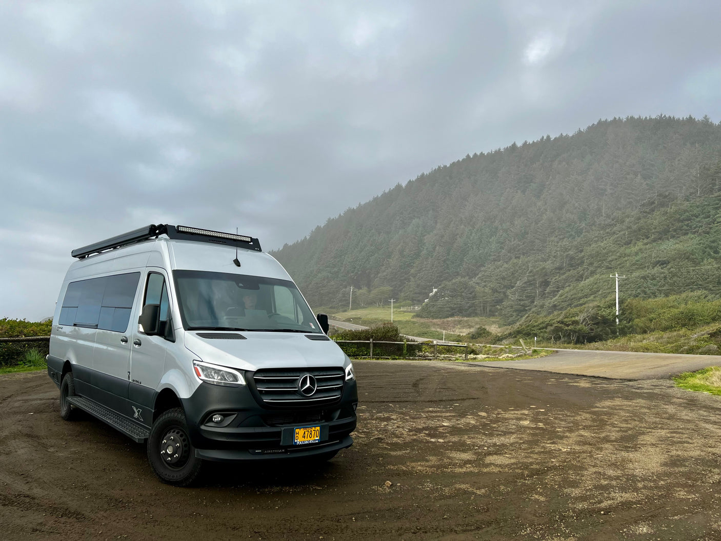 Silver Mercedes camper van RV rental parked on a dirt lot, with a misty forest and road in the background.