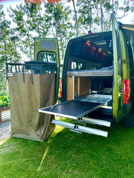 Green RV rental with open rear doors showing a bed & slide-out tray, plus an attached privacy tent in a grassy outdoor setting.