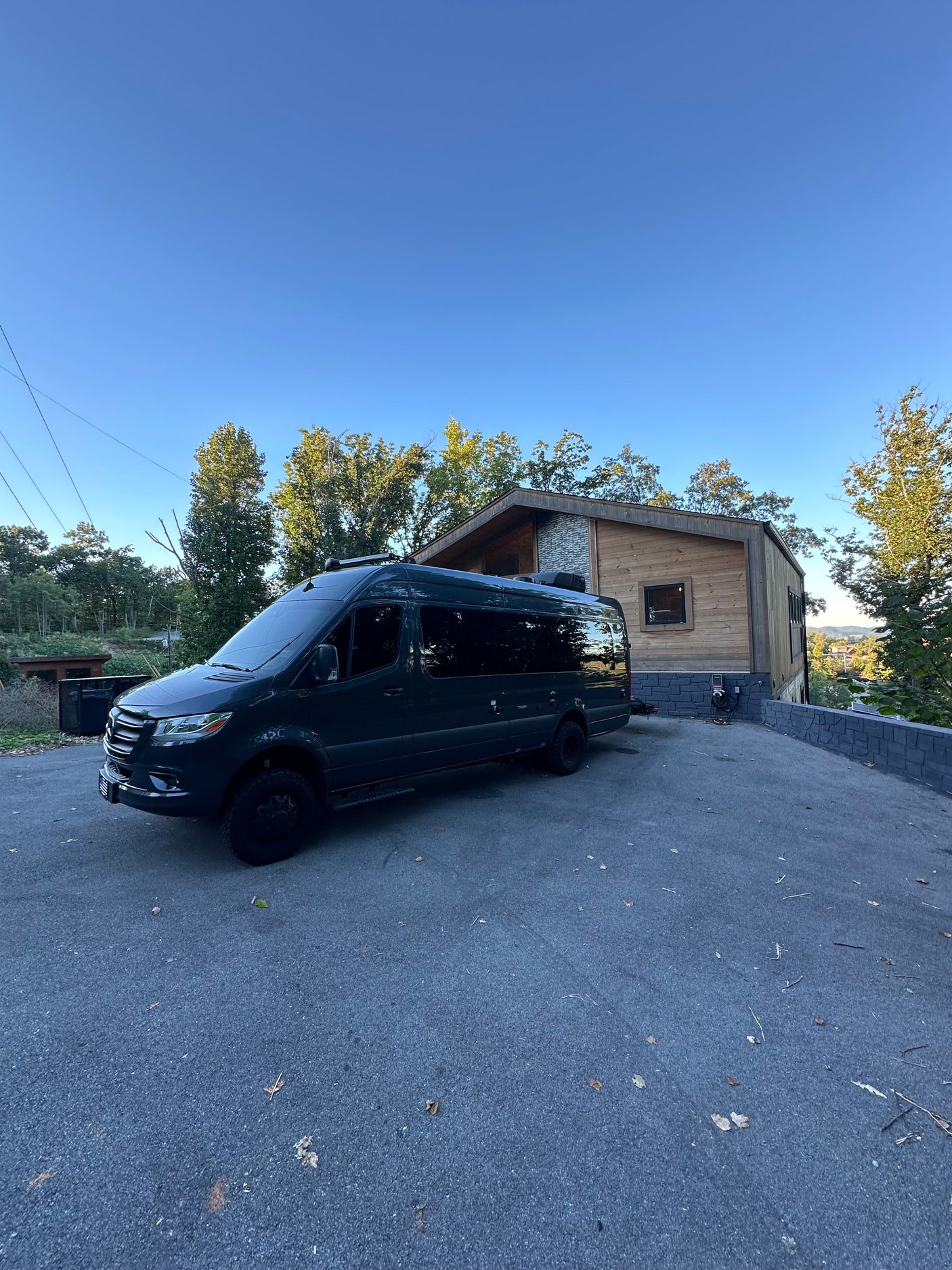 Dark Mercedes Sprinter van (RV rental) parked on a sunny day outside a modern house with trees.