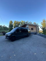 Dark Mercedes Sprinter van (RV rental) parked on a sunny day outside a modern house with trees.