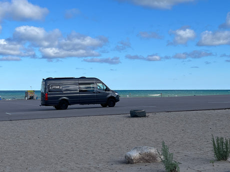 A dark grey RV rental is parked on an asphalt lot by a sandy beach with blue water and clouds.