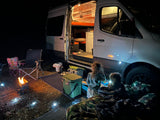 Cozy night camping in an RV rental! Kids playing outside a lit camper van with a fire pit & string lights.