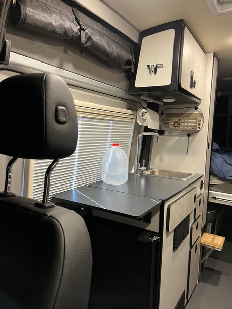 This RV rental features a compact kitchen with a sink, counter, and fridge. Overhead storage & window blinds visible. Ideal for travel.