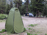 A green privacy tent and an RV rental teardrop trailer are set up at a serene mountain campsite, with a person near the trailer.
