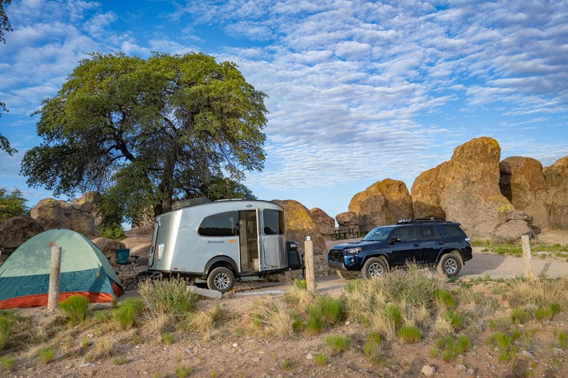 RV rental adventure! A small silver travel trailer and blue SUV parked at a rocky, tree-filled campsite with a tent and big sky. — photo 12