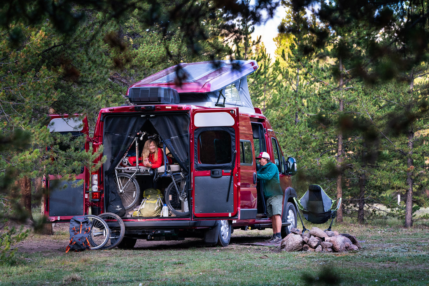 This RV rental features a red pop-top camper van in a forest campsite. A couple and their bikes are visible, enjoying the outdoors.