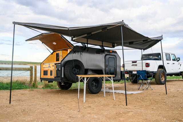 This XTR teardrop **rv rental** and Jeep Gladiator create a perfect lakeside overlanding camp, with an awning, table, and chair.
