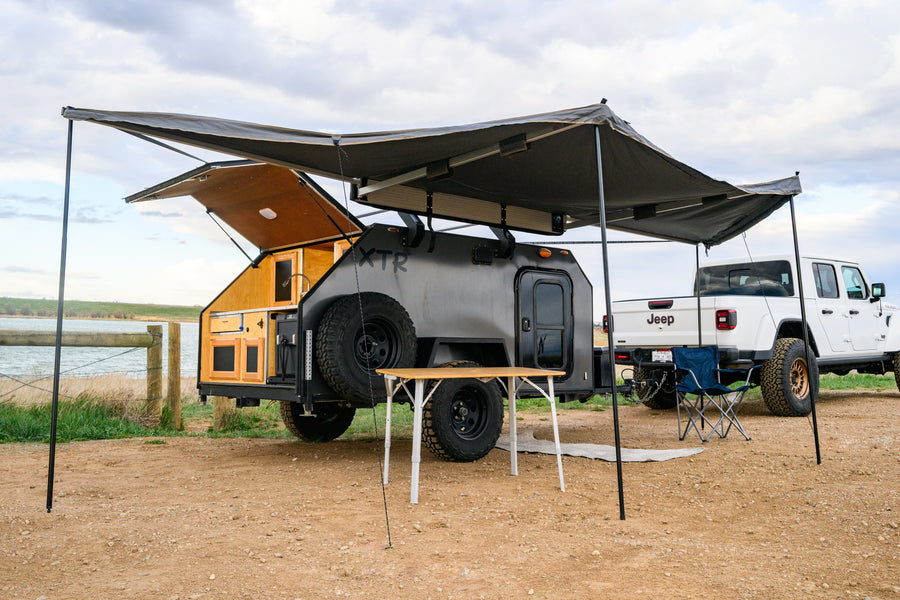 This XTR teardrop **rv rental** and Jeep Gladiator create a perfect lakeside overlanding camp, with an awning, table, and chair.