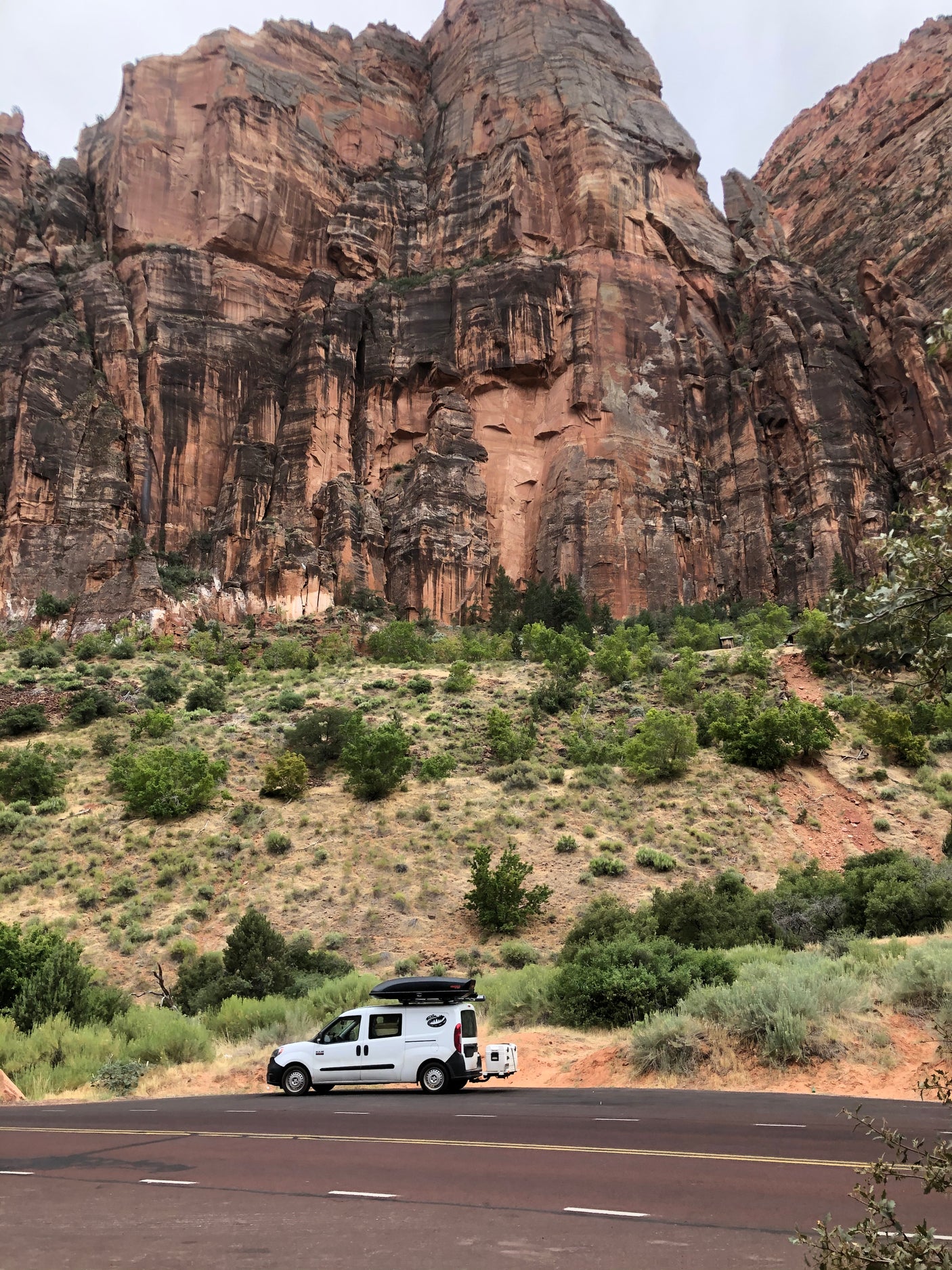 An RV rental camper van pauses on a road, framed by towering red rock cliffs and a green, arid hillside. Ideal for national park adventures.