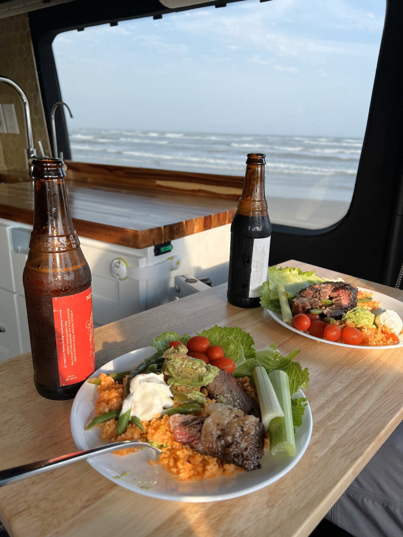 Beachfront RV rental dining: two plates of food, beer, and a serene ocean view from inside a van.
