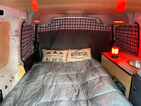 An RV rental's cozy interior, showing a bed with themed pillows, plaid curtains, a sink, and accent lighting.