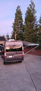 RV rental van with illuminated awning and flag. Sunset reflects in windows; parked on driveway. Ready for adventure.