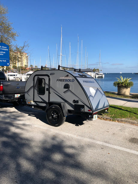Grey Freesolo teardrop rv rental hitched to a truck, parked by a marina filled with sailboats. Ideal for outdoor adventures.