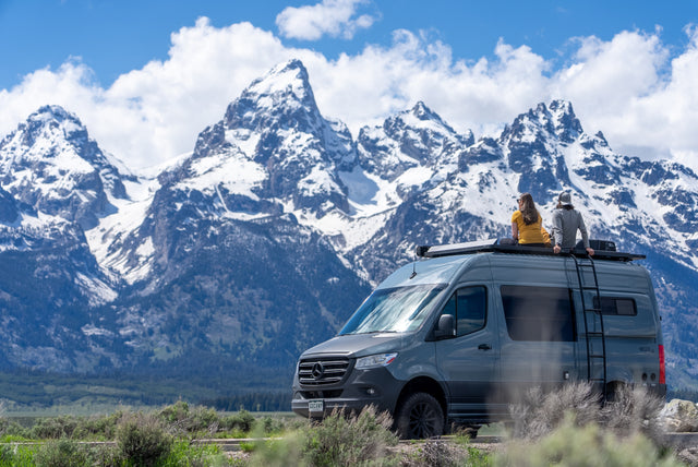 Couple on an RV rental enjoys majestic snow-capped mountains from their camper van roof.