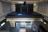 RV rental interior: A bed/lounge area with patterned gray cushions and a dining table on the right.