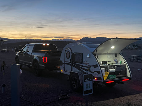 A pick-up truck with an RV rental at a campsite under a serene sunset, kitchen open and lit. Mountains silhouette the horizon.