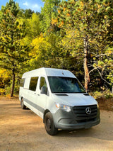 White Mercedes Sprinter camper van, ideal for rv rental adventures, parked on a dirt path amidst vibrant autumn trees.