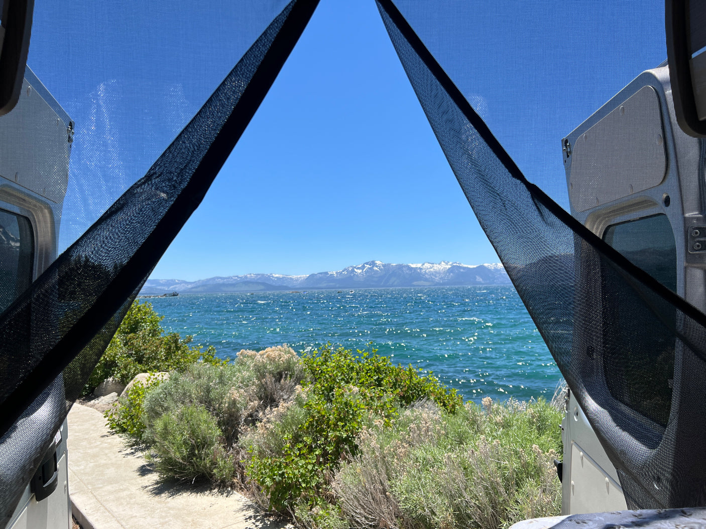 Stunning lake view from an RV rental, framed by screens. Snowy mountains across sparkling water and green plants.