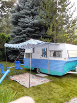 This colorful teal & blue RV rental is set up for lake house camping, with an awning, chairs, and rug on the grass.
