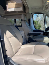 Front cabin of an RV rental with comfortable, light-colored captain's chairs, overhead storage, and a bright outdoor view.